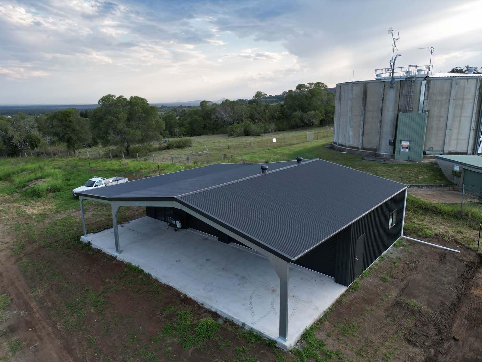 Dark steel shed with a skillion awning on a concrete slab, rural hills and water tanks behind